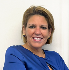 A woman with short blonde hair wearing a blue blazer, smiling, against a white wall background.