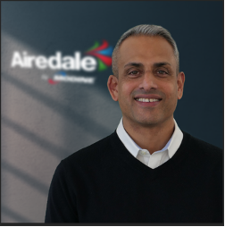 A man with short black hair, wearing a black sweater and white collared shirt, smiling in front of a gray wall with the Airedale logo.