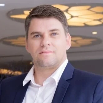 Headshot of a man in a navy blazer and white shirt, indoors with decorative lighting in the background.