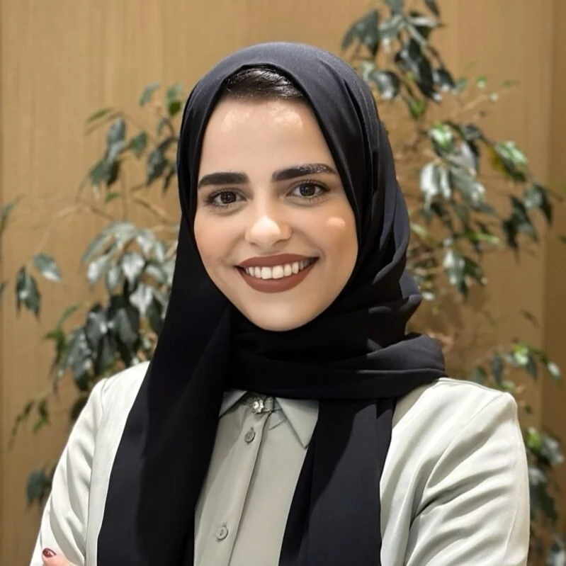 A young woman with dark hair and brown eyes, smiling, wearing a black hijab and a light-colored blouse with a brooch at the collar, posing in front of a plant on a wooden background.