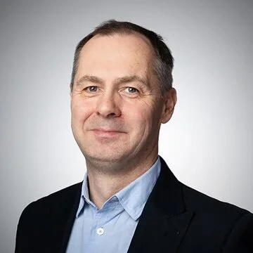 Professional headshot of a middle-aged man with short dark hair, wearing a light blue collared shirt and a dark blazer, smiling slightly against a gray background.