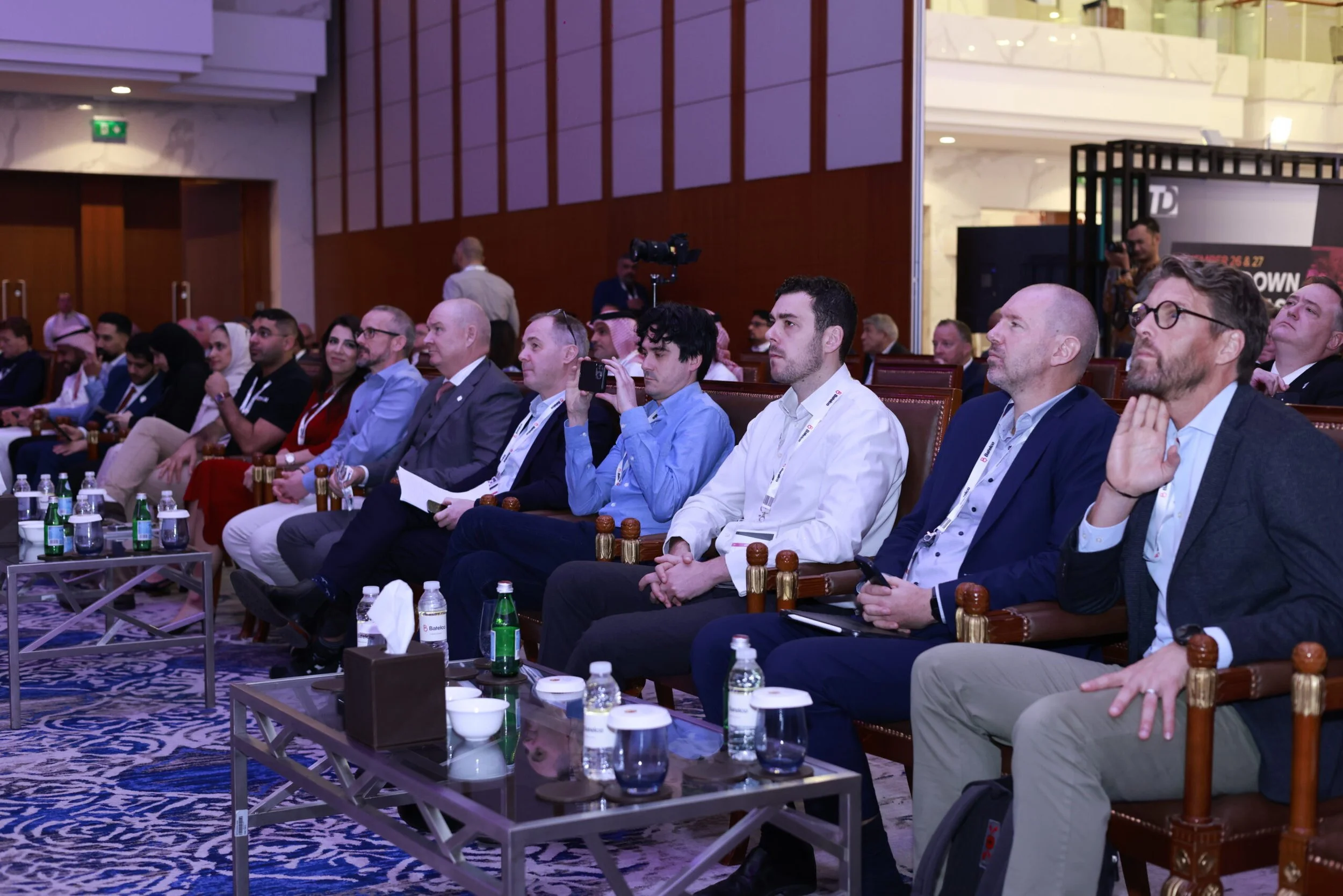 People attending a conference or seminar sitting in chairs facing forward, with some holding phones, in a room with marble walls and wooden panels.