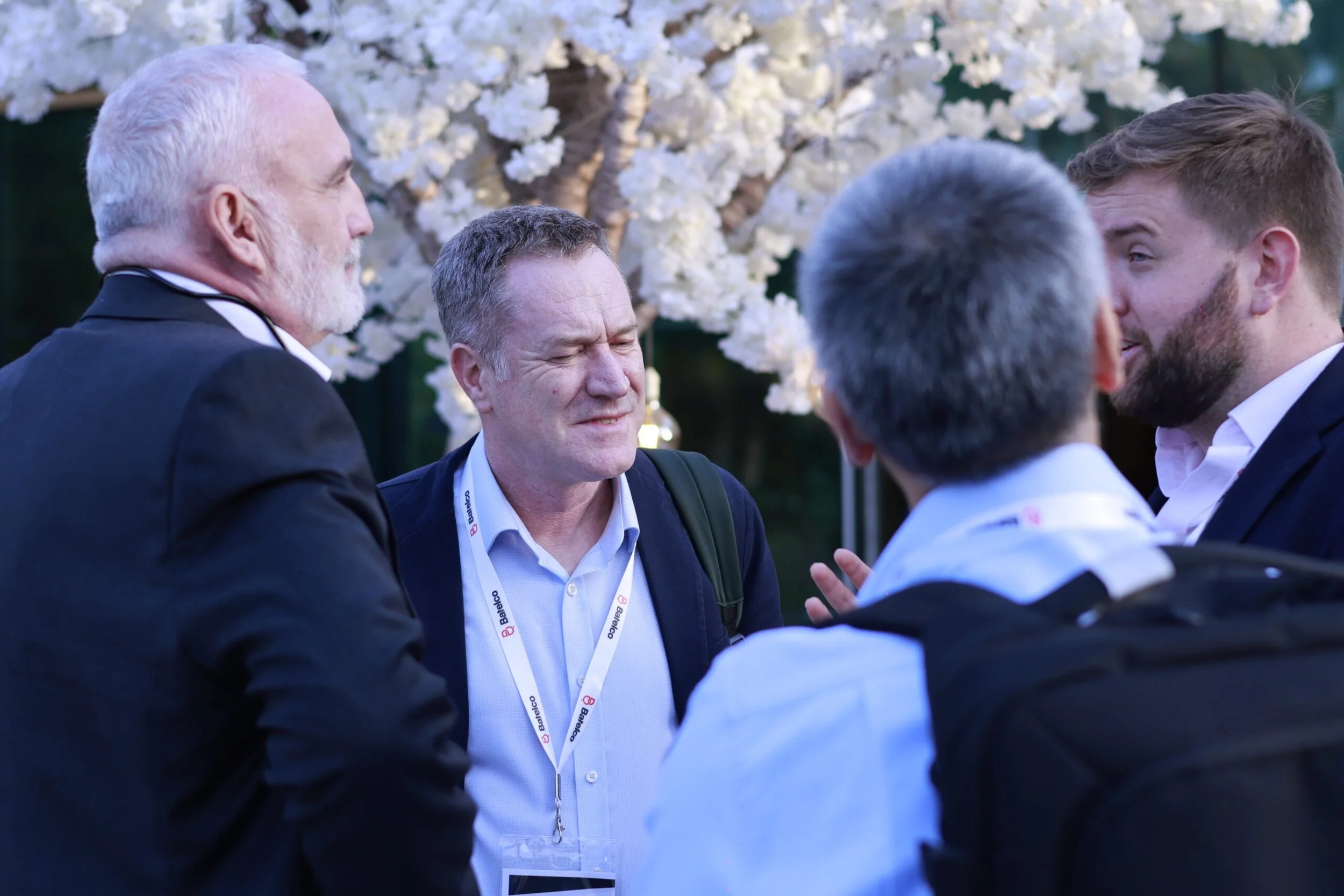 Group of people engaged in conversation outdoors, with a background of white flowers on a tree.