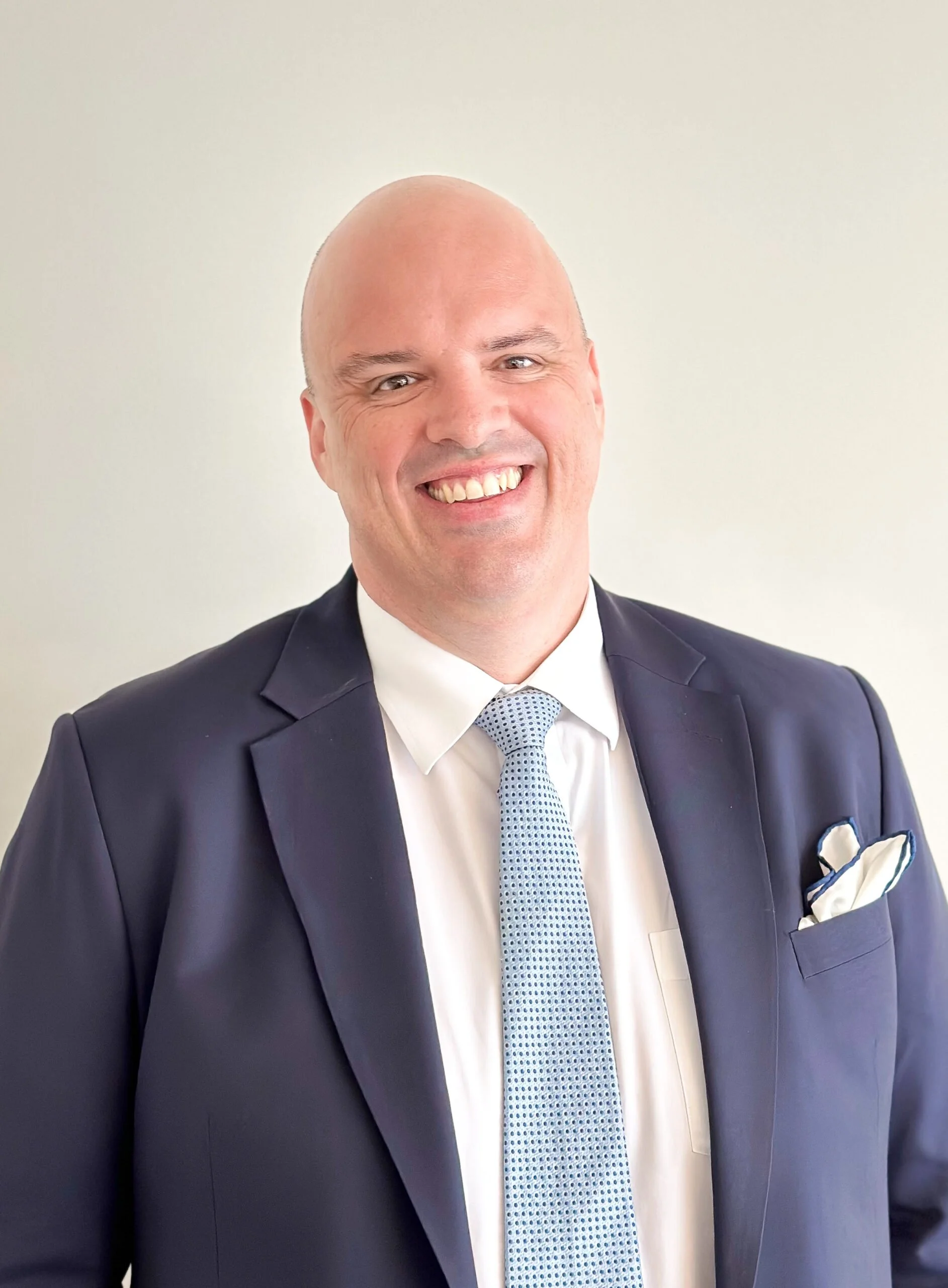 A man in formal business attire smiling, with a white shirt, blue patterned tie, and dark suit jacket with a pocket square, standing against a plain light-colored background.