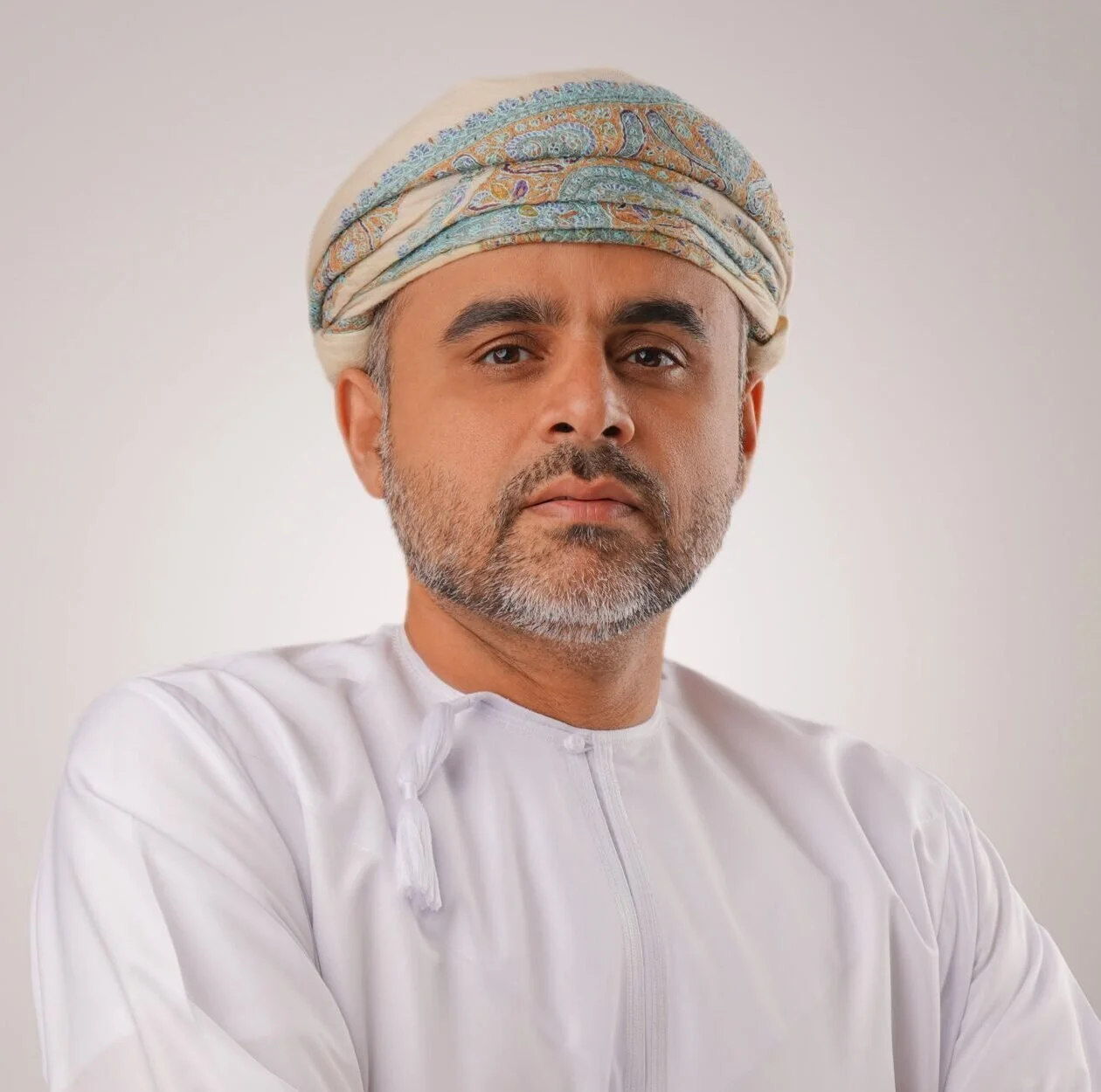 A middle-aged man wearing a white traditional Middle Eastern kandura and a colorful turban, posing against a plain background.