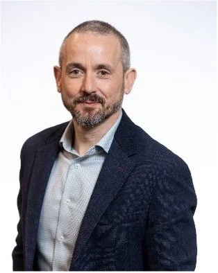 A man with short hair and a beard wearing a dark blazer and light-colored shirt, smiling against a white background.