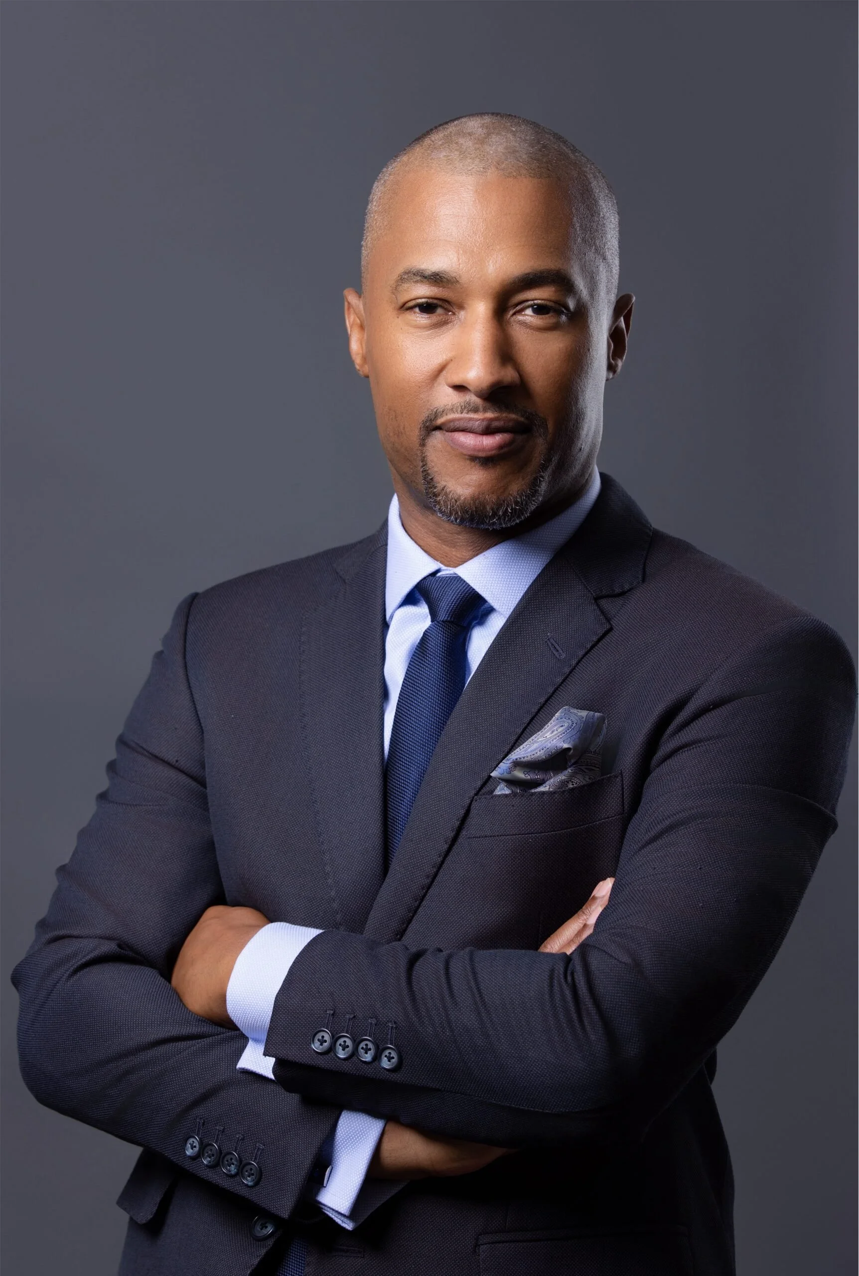A confident African American man in a dark suit, white shirt, and blue tie, standing with arms crossed against a gray background.