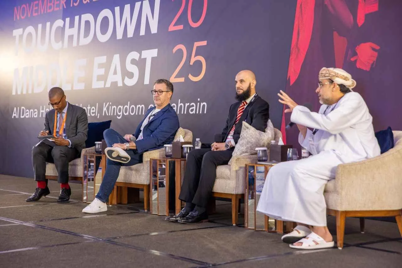 A panel of four men seated on stage at a conference, with a large backdrop behind them displaying event details in white and pink text and a logo. The men are dressed in formal and traditional attire, engaged in discussion during the event.