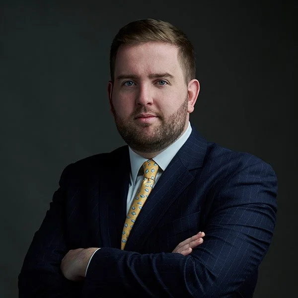 A man with a beard and short hair in a dark suit with a yellow tie, posing with crossed arms against a dark background.