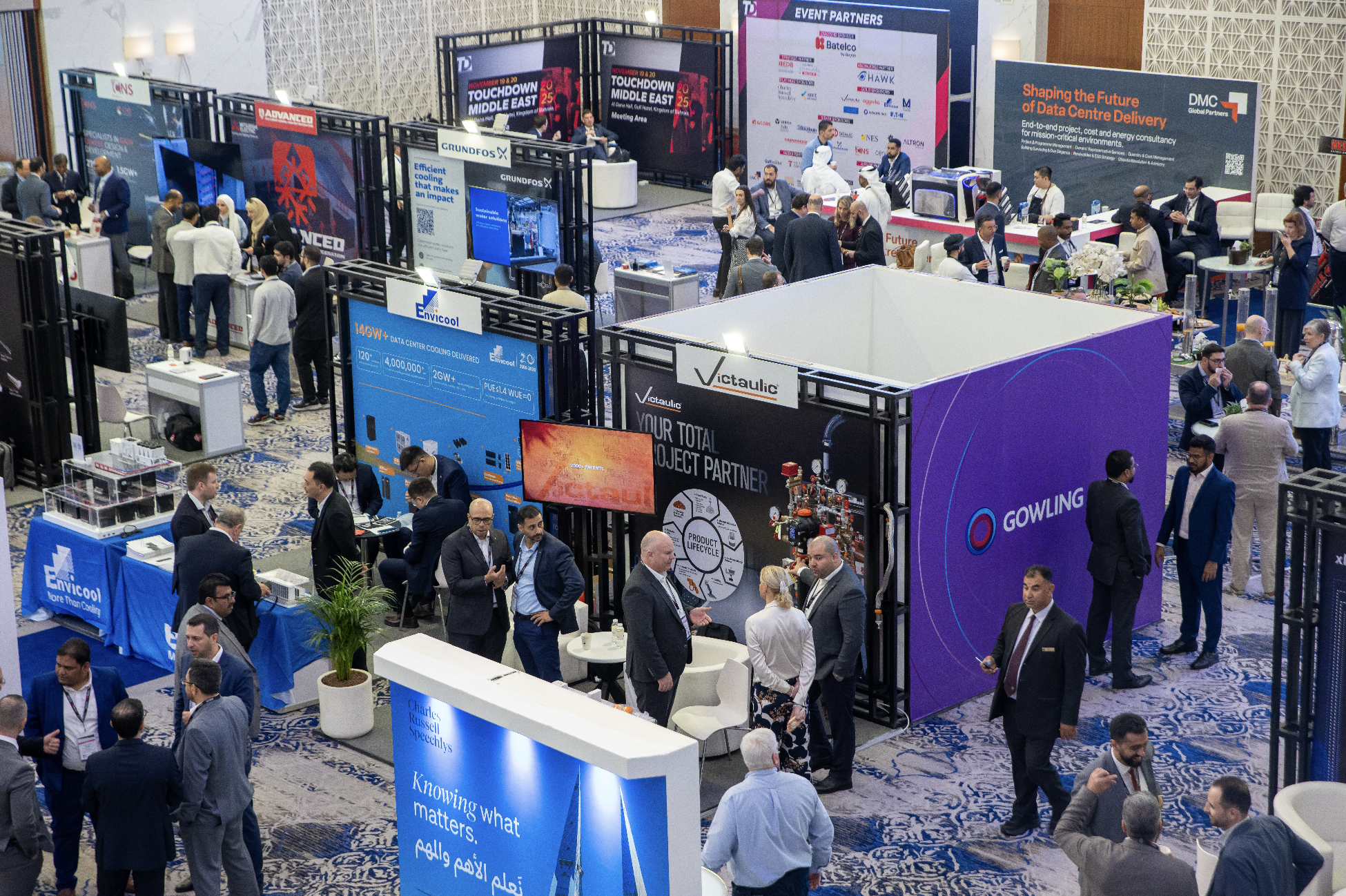 A busy conference or trade show with various exhibition booths, people networking, and standing around discussing products or services. The floor is carpeted with a patterned blue and beige rug, and there are several potted plants and informational display screens visible.