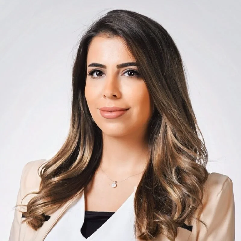 A young woman with long, wavy brown hair, wearing a beige blazer and a delicate silver necklace, smiling slightly against a neutral background.