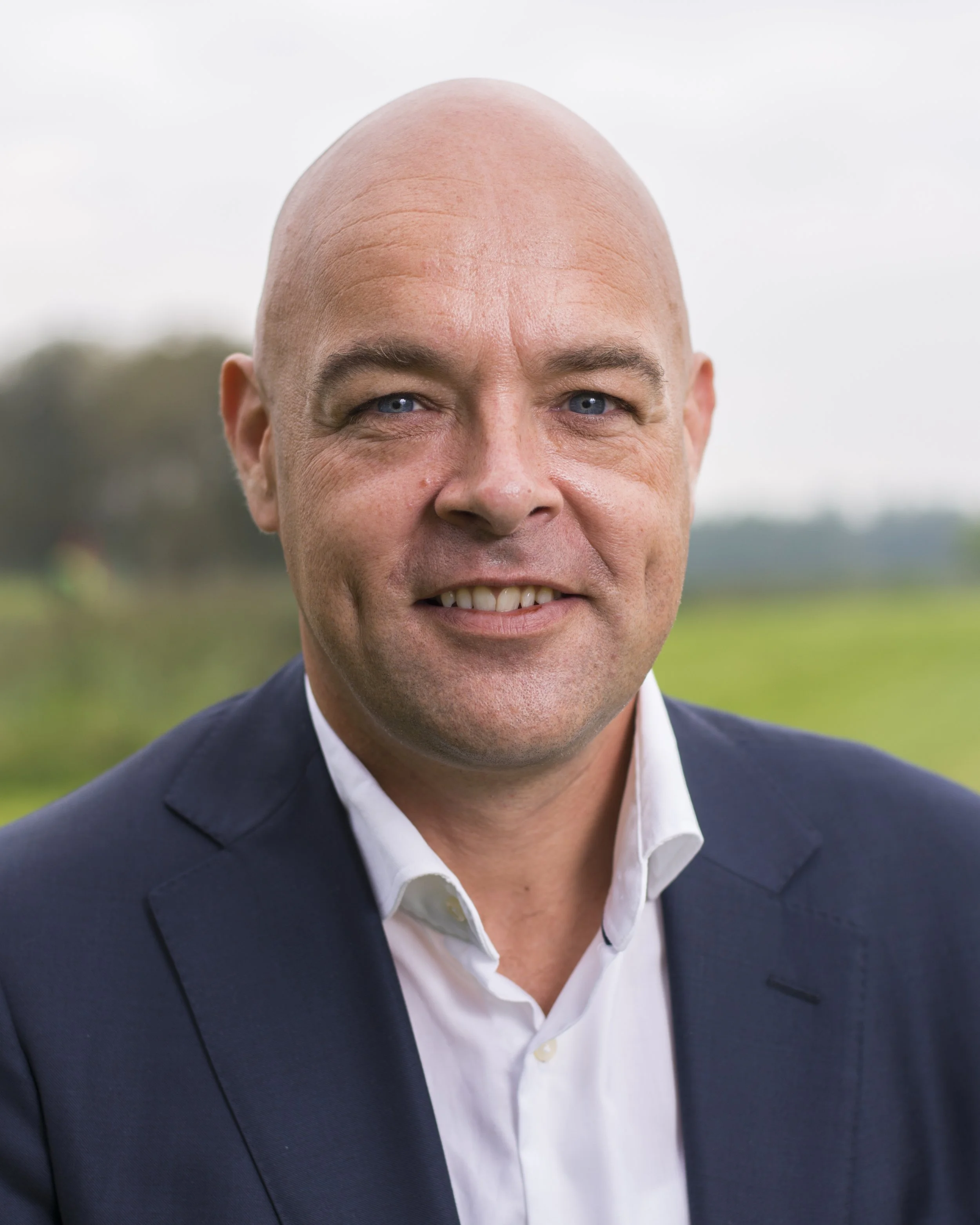 Close-up of a smiling middle-aged man in a navy blazer and white shirt, standing outdoors with a blurred background of greenery and cloudy sky.