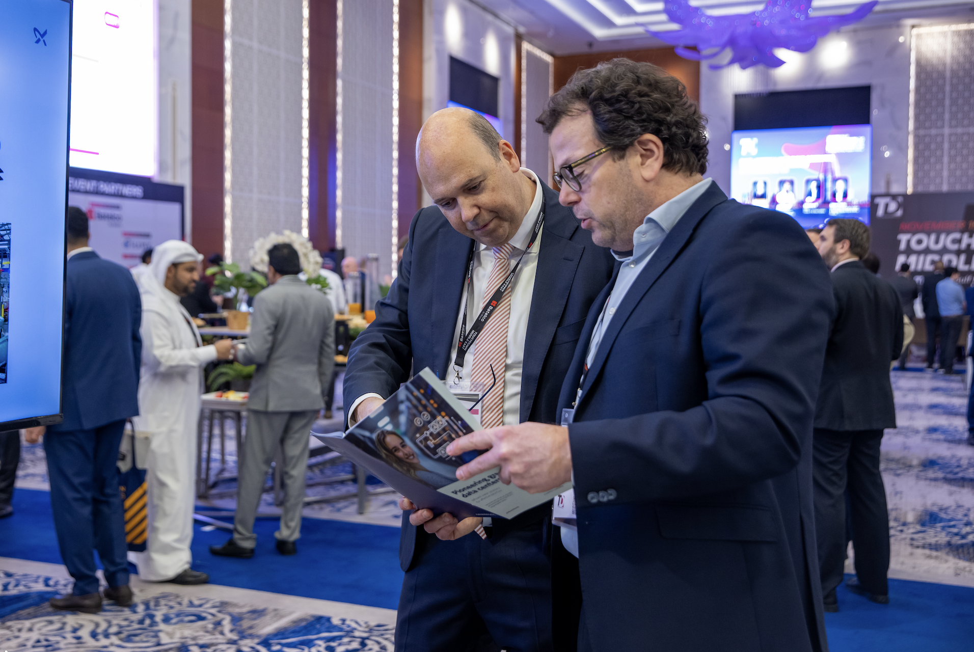 Two men in suits looking at a brochure at a professional conference or event in a large, decorated conference hall with screens and other attendees in the background.