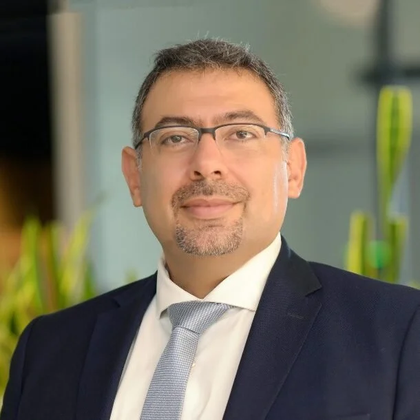 A man in a business suit with glasses, short hair, and facial hair, smiling in an indoor setting with plants in the background.