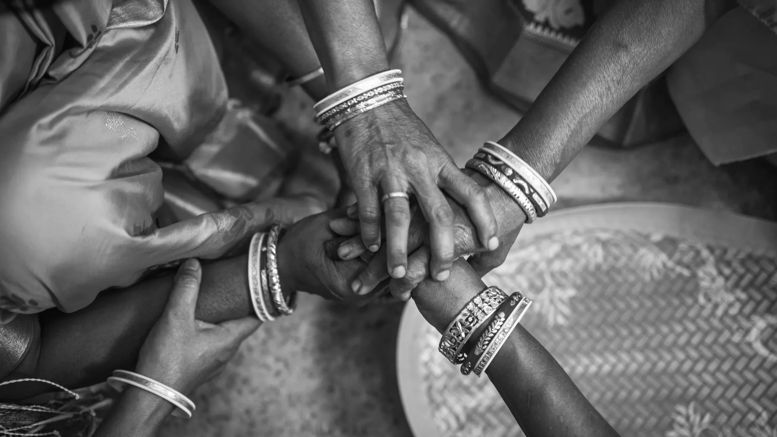Black and white photograph of multiple hands stacking together, with each hand wearing various bracelets and rings, symbolizing unity and teamwork.