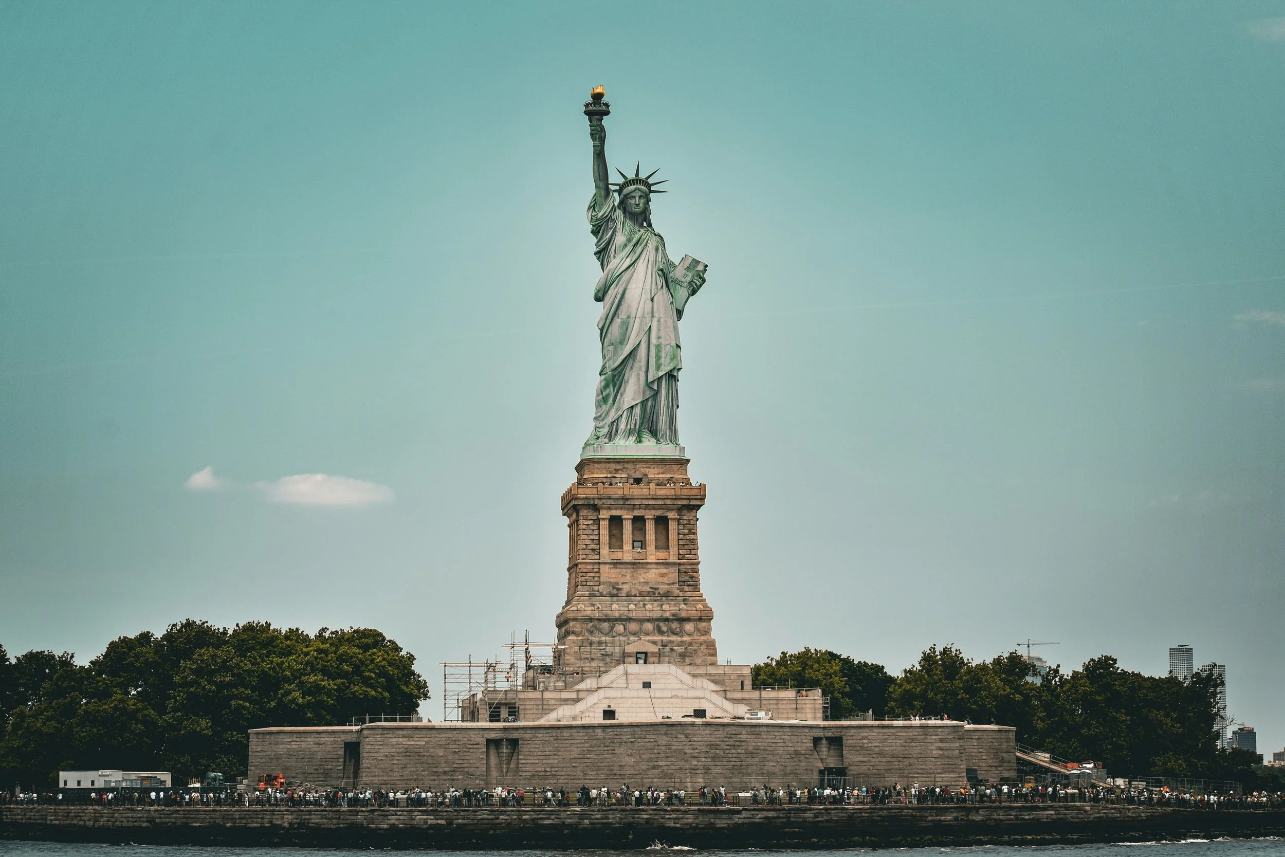 The Statue of Liberty stands tall on Liberty Island with a clear blue sky and a few clouds behind it. The statue holds a torch and a tablet, symbolizing freedom and independence. There is a group of visitors at the base, and trees and city buildings are visible in the background.