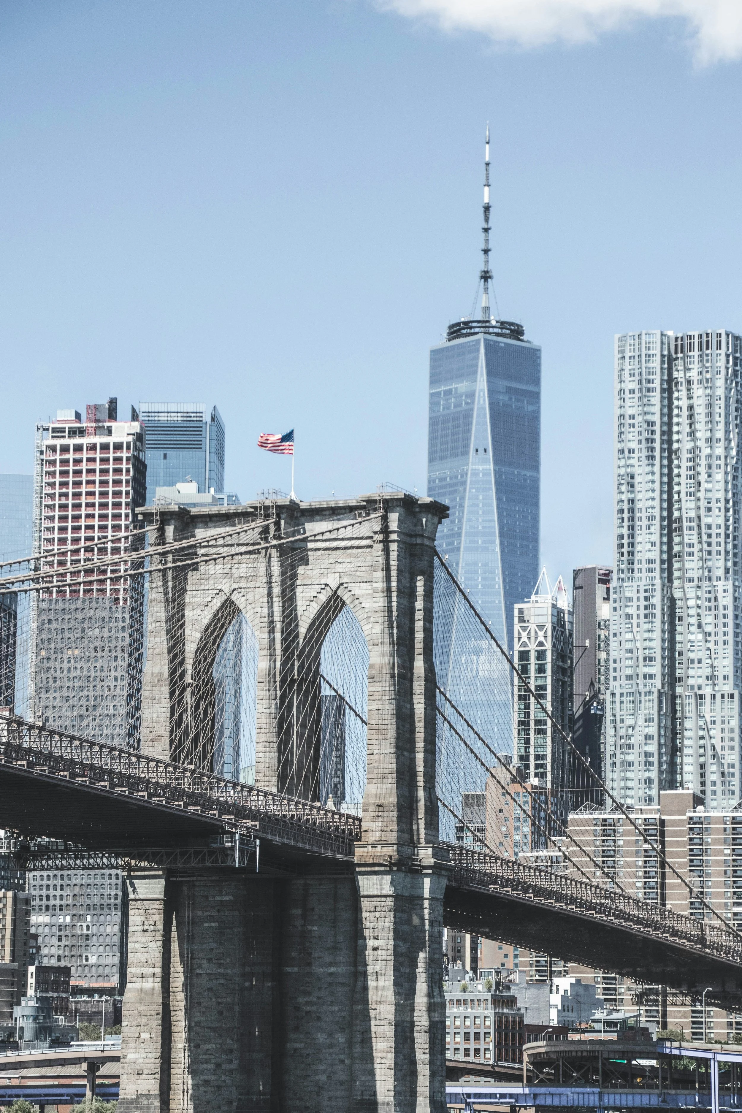 View of the Brooklyn Bridge with the Manhattan skyline in the background, including One World Trade Center, a telecommunications tower, and modern high-rise buildings.