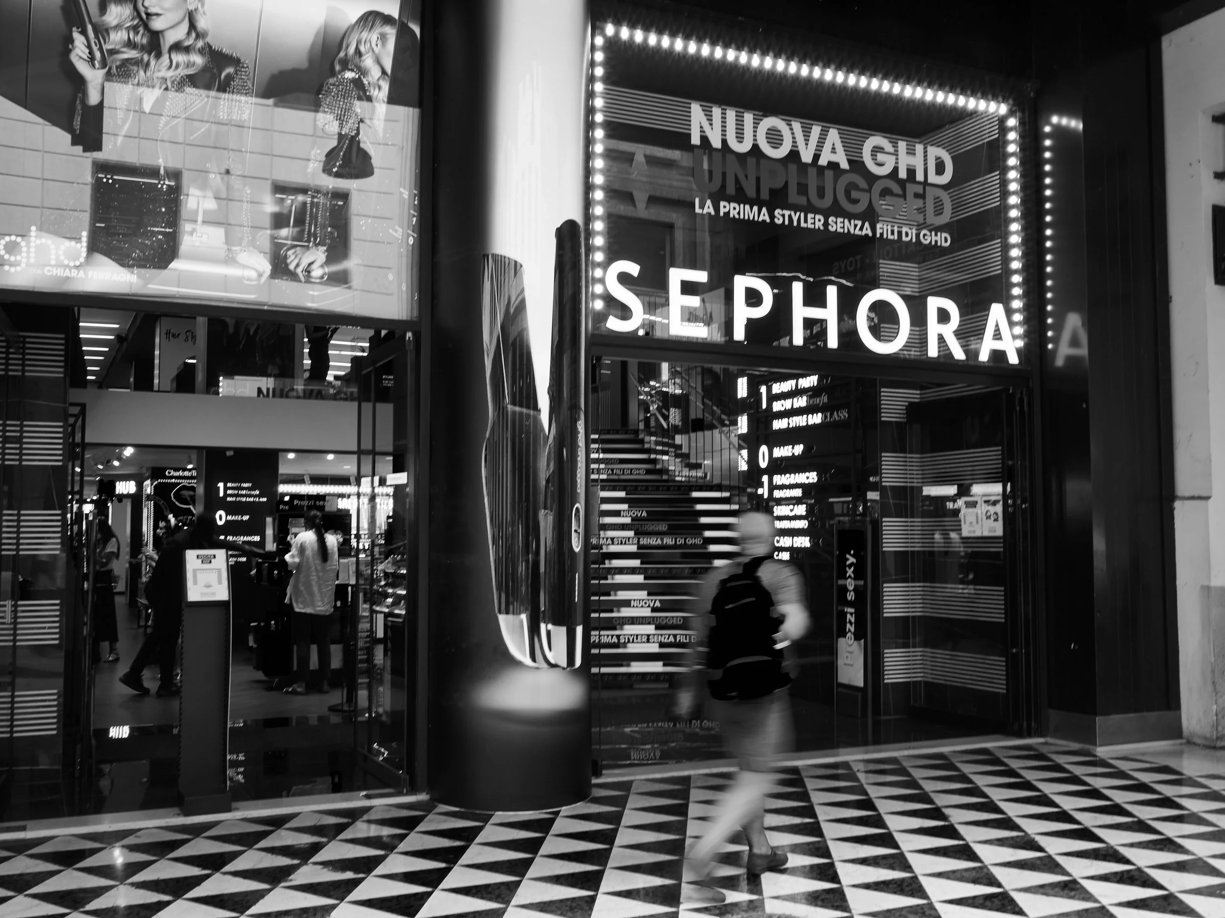 International shopping mall entrance with bright signage for Sephora and other beauty stores, with people walking inside, black and white photo.