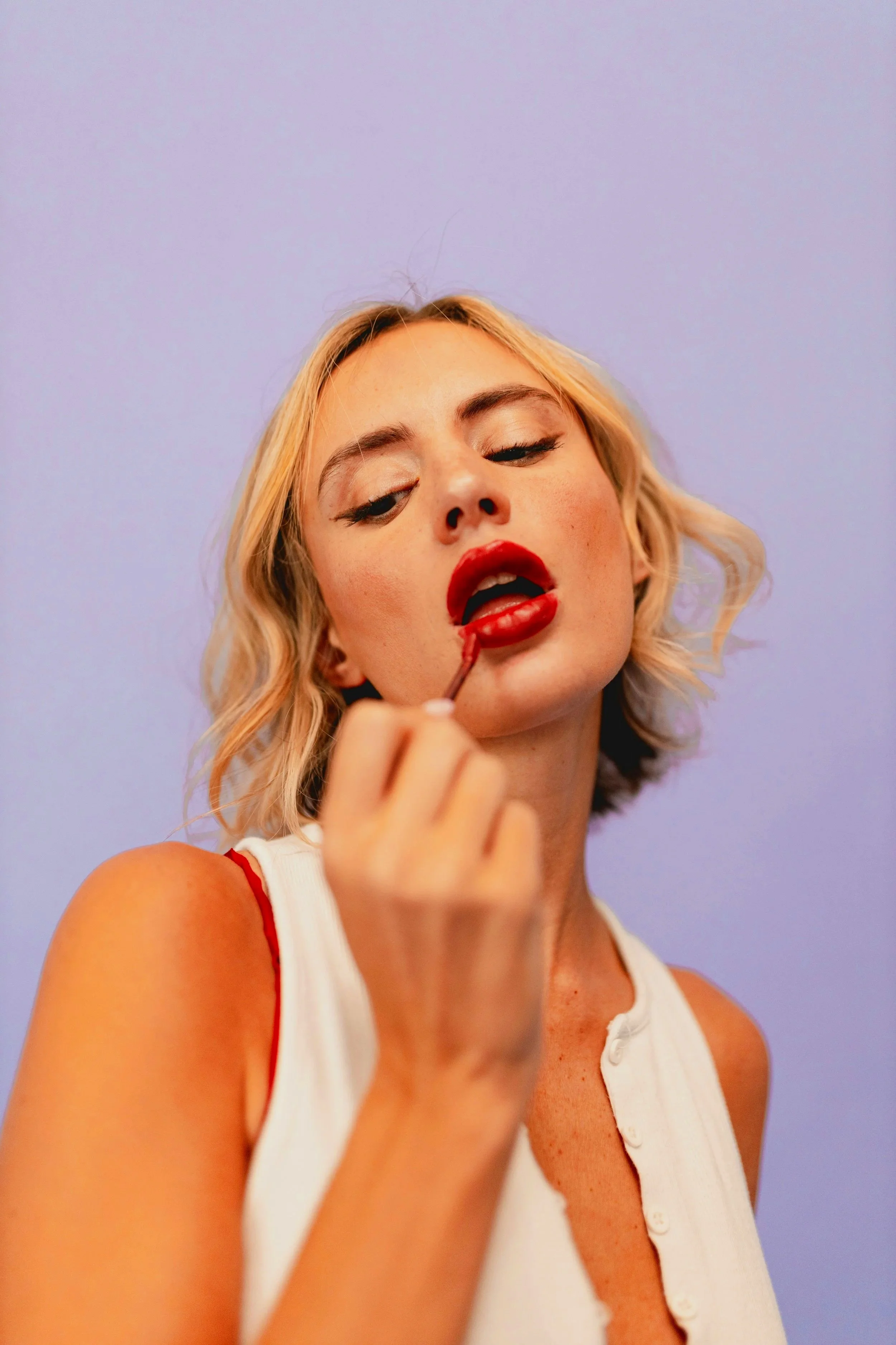 Close-up of a woman with short blonde hair, red lipstick, and wearing a white top with red straps, applying lipstick against a purple background.