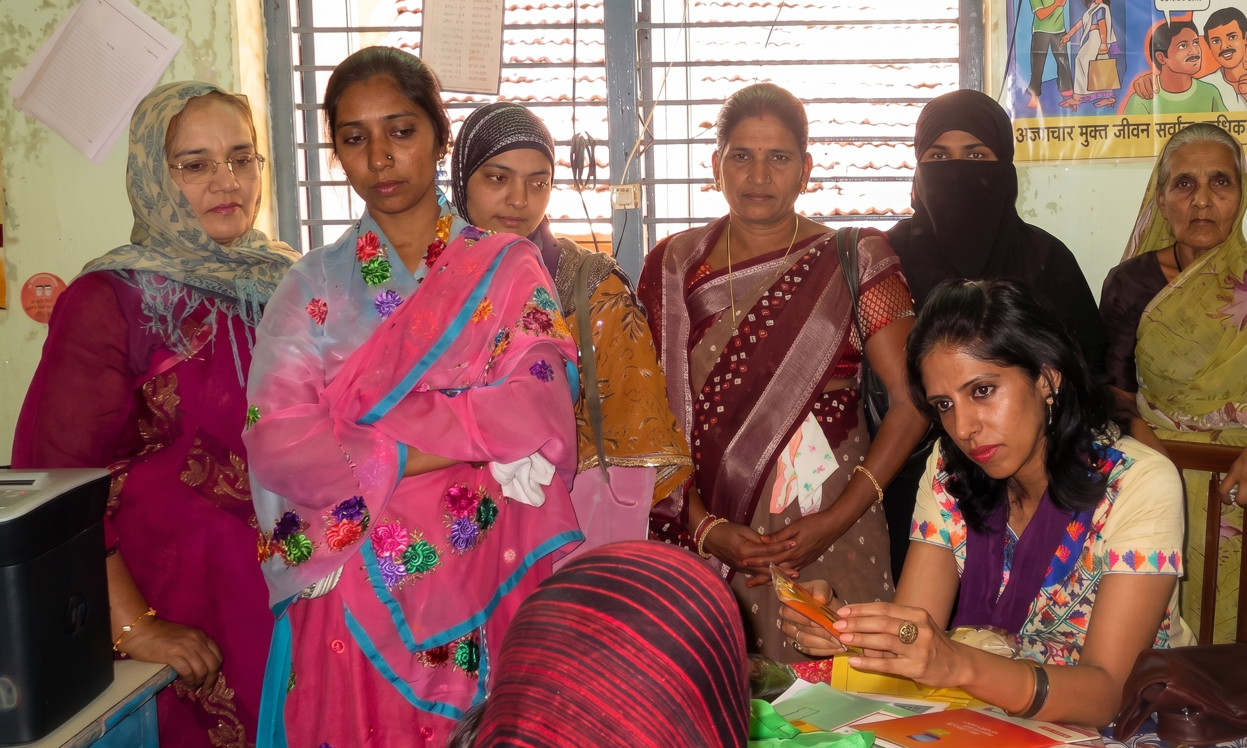 A group of women, some wearing sarees and others in traditional attire, gathered indoors near a window with metal bars. One woman is seated at a desk, holding a yellow card, while others stand around her, observing. The scene appears to be in a community or administrative setting, with a colorful poster on the wall and documents on the table.