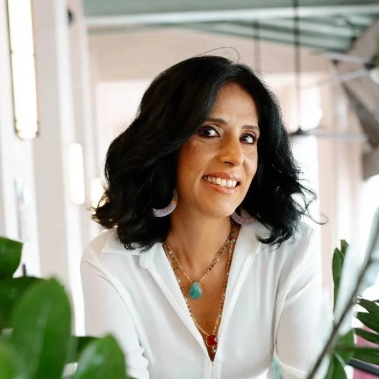 A woman with black hair, wearing a white shirt and colorful jewelry, smiling in an indoor setting with plants.