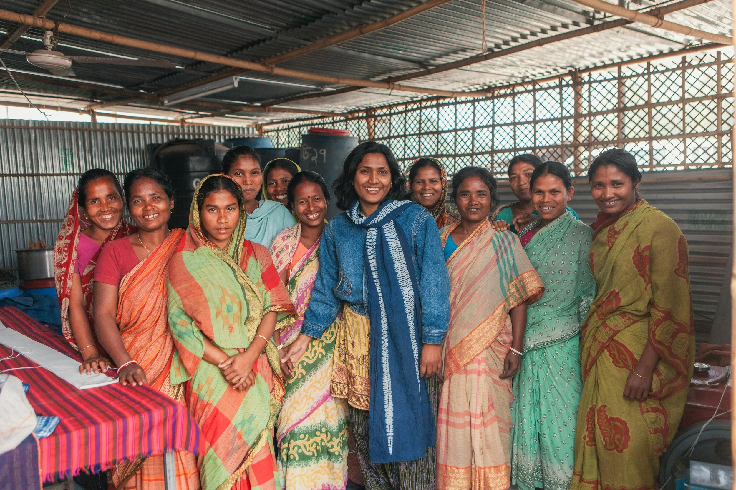A group of women dressed in colorful traditional Indian attire, smiling, standing inside a metal structure with a corrugated roof.