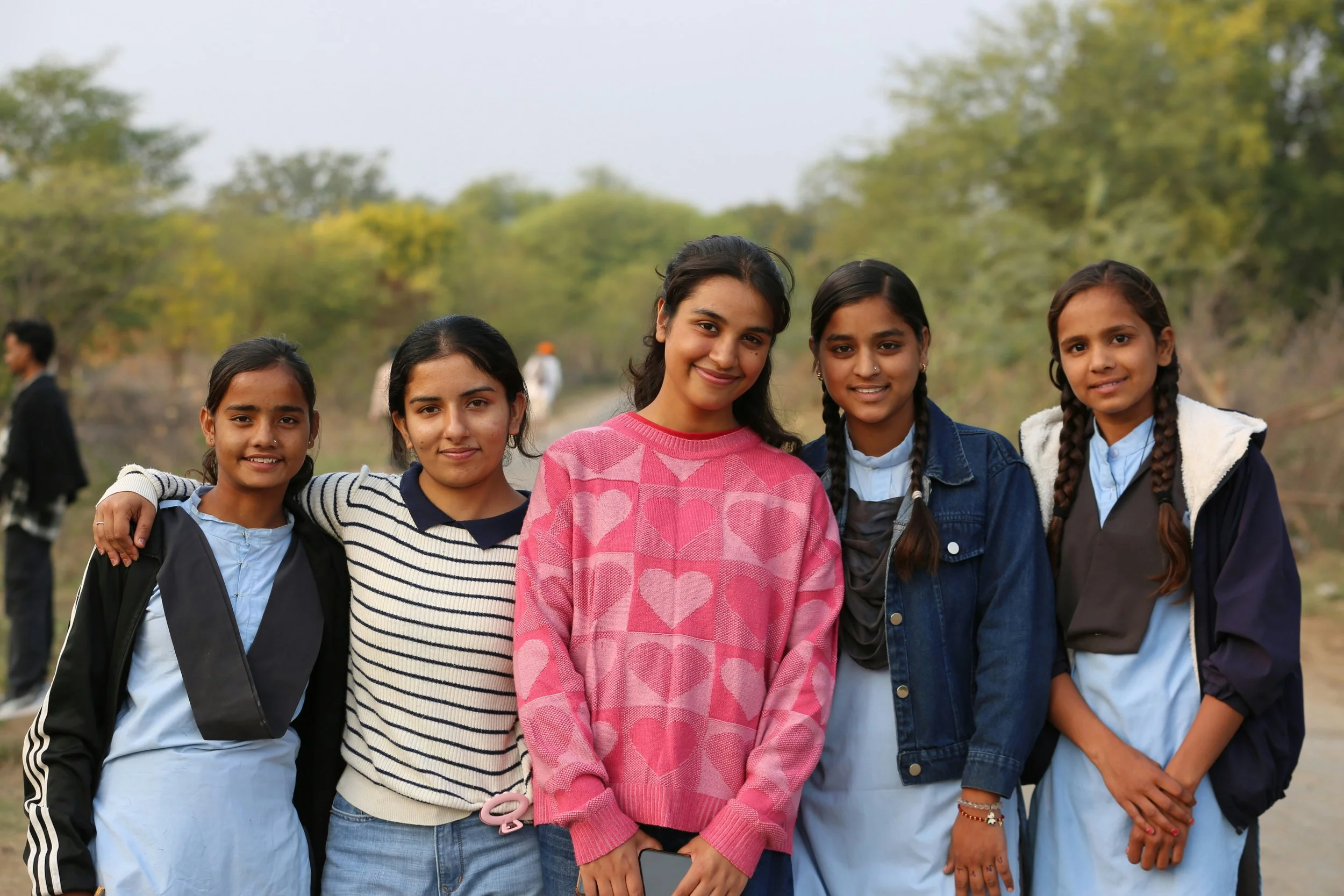 Five young girls standing outdoors close together, smiling at the camera, with trees and other people in the background.