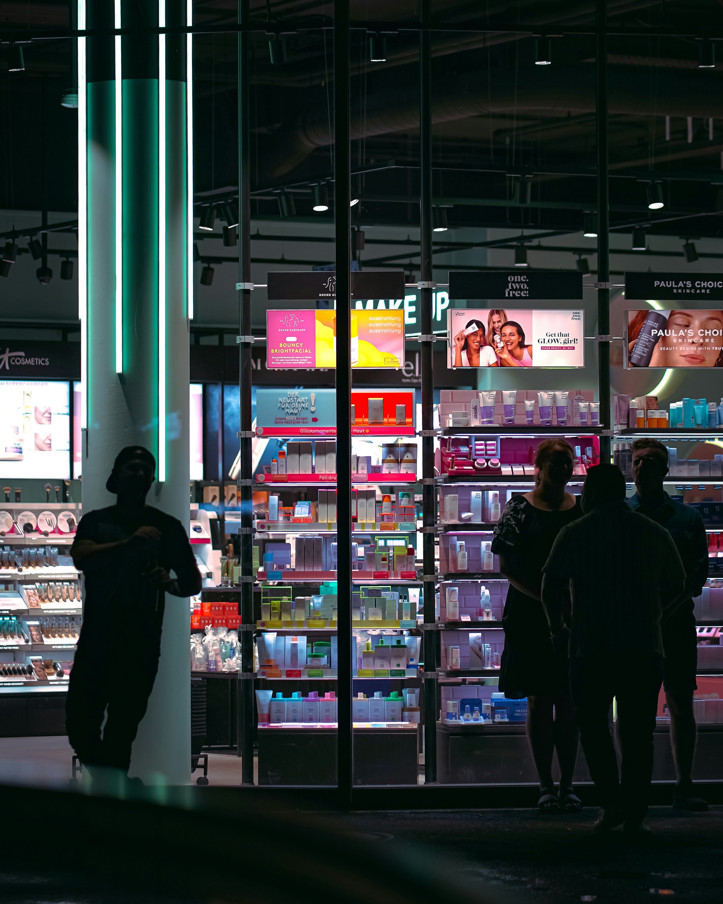 People shopping at a store counter inside a mall, viewed through large glass windows at night.