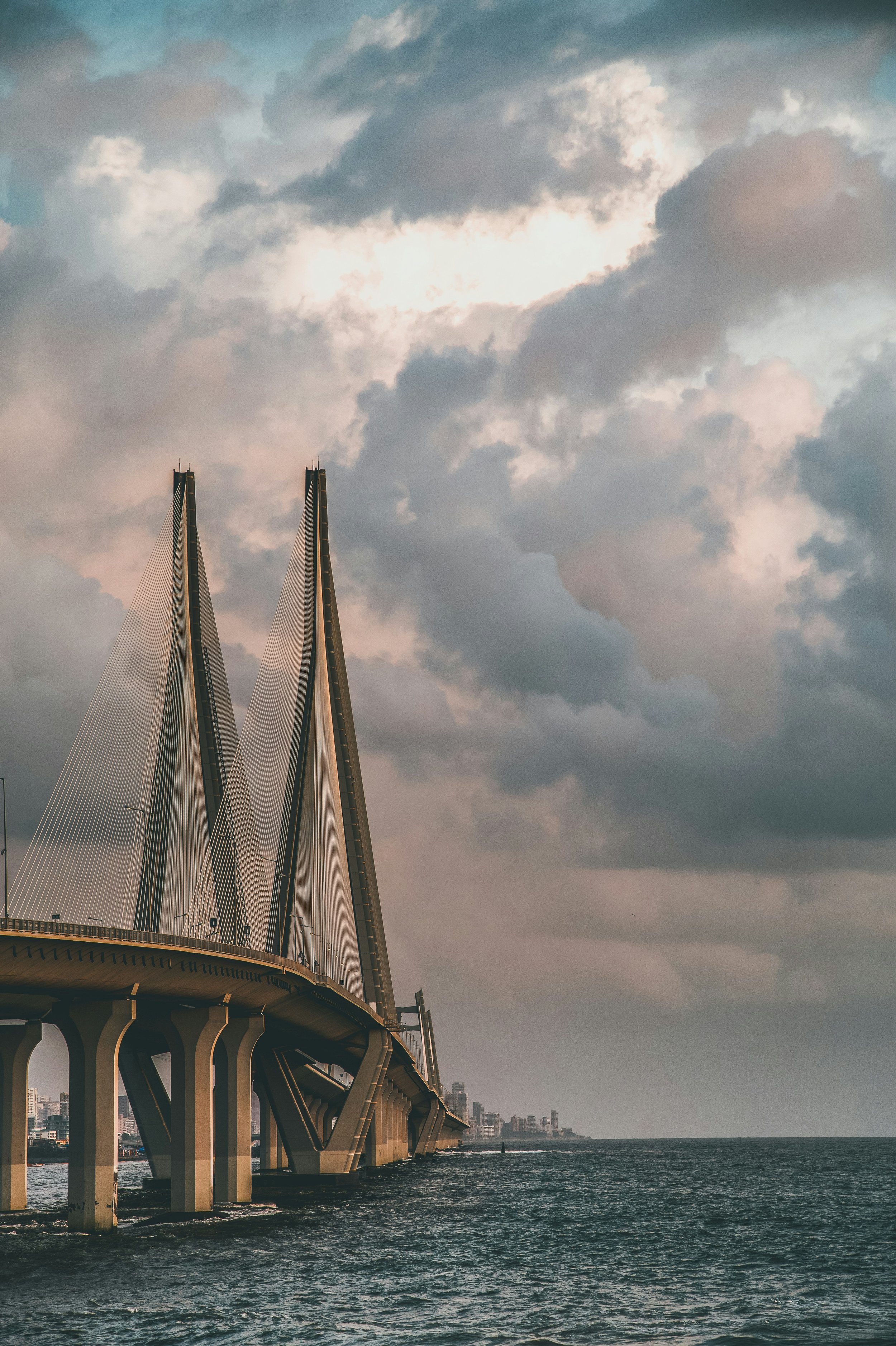 A bridge with tall, cable-stayed towers extending over a body of water, with a cloudy sky overhead and a distant city skyline in the background.