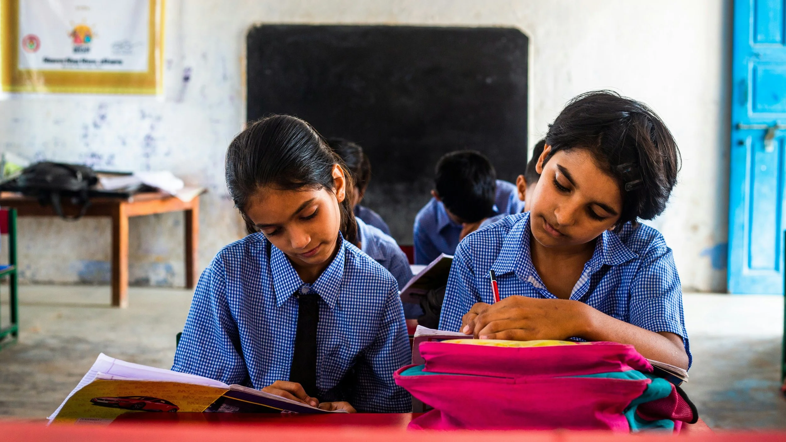 Two schoolgirls in school uniforms sitting at a desk, focused on writing or reading, with backpacks on the desk and other students and a blackboard in the background.