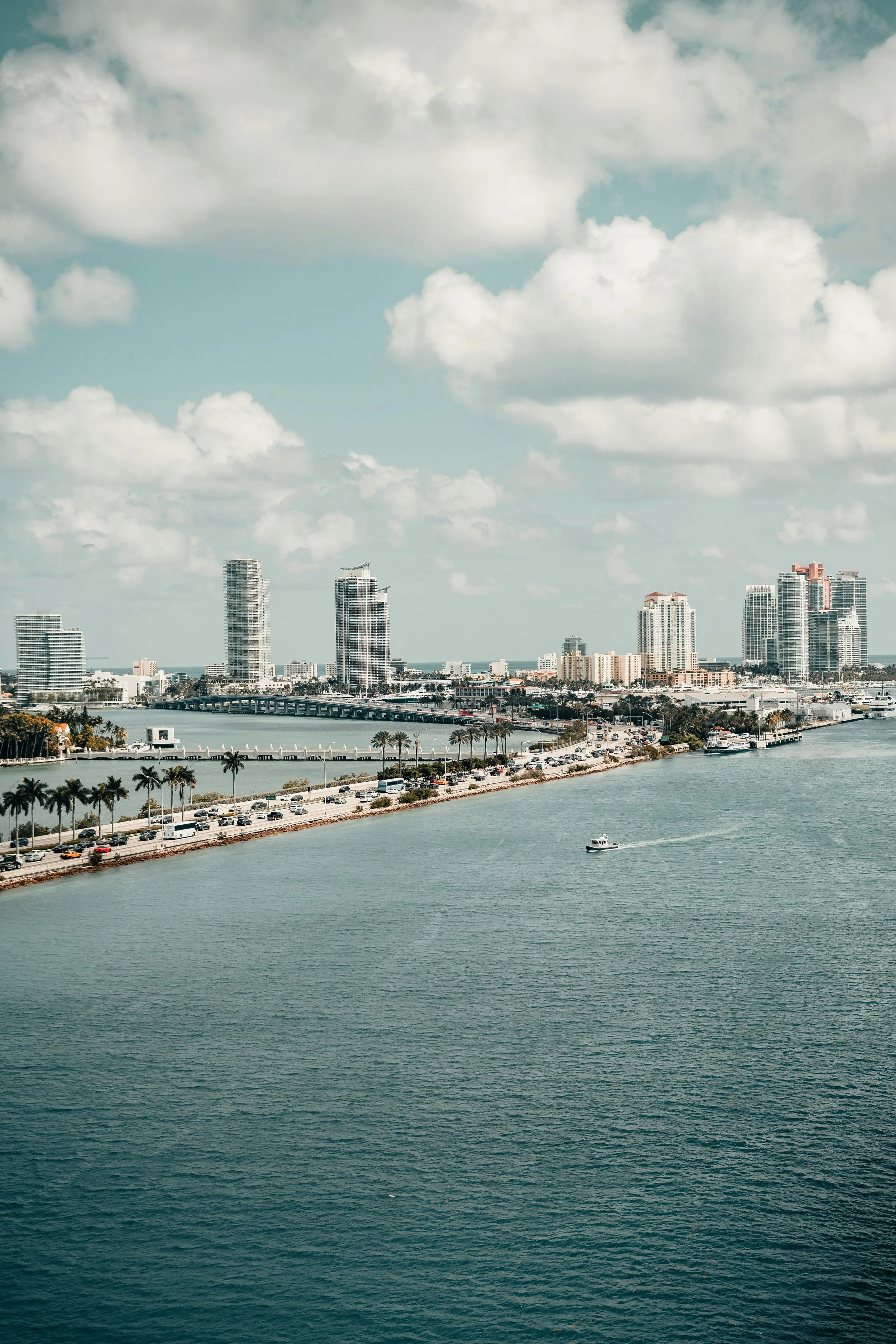 Skyline of a coastal city with high-rise buildings, a bridge, and water with a boat, under a partly cloudy sky.