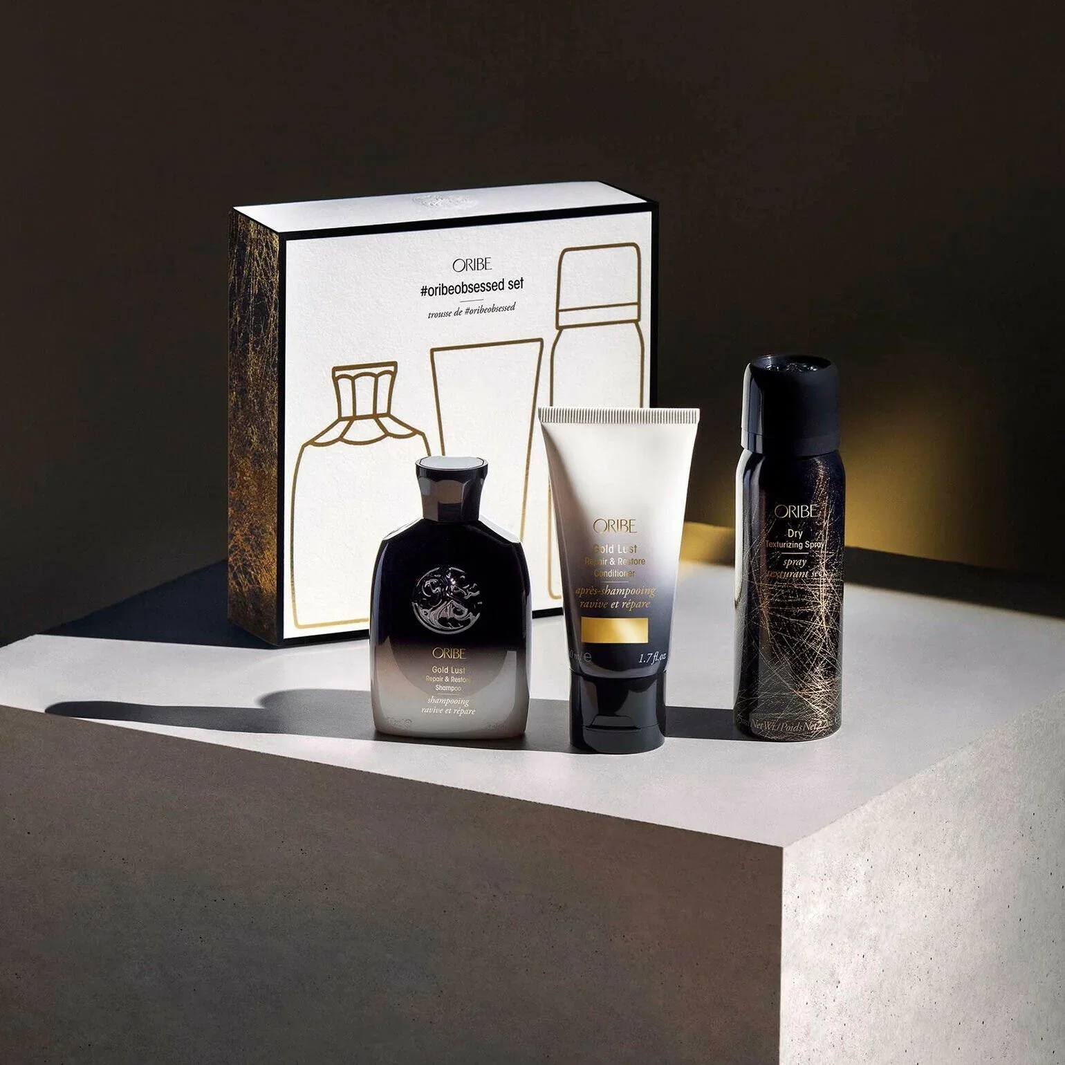 Set of Oribe hair care products including a shampoo, conditioner, and spray, displayed on a white surface with a patterned box behind them.