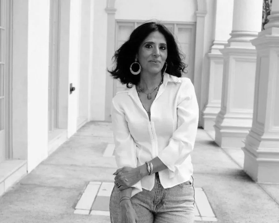 A woman with dark hair wearing a white blouse and jeans, standing indoors near architectural columns.