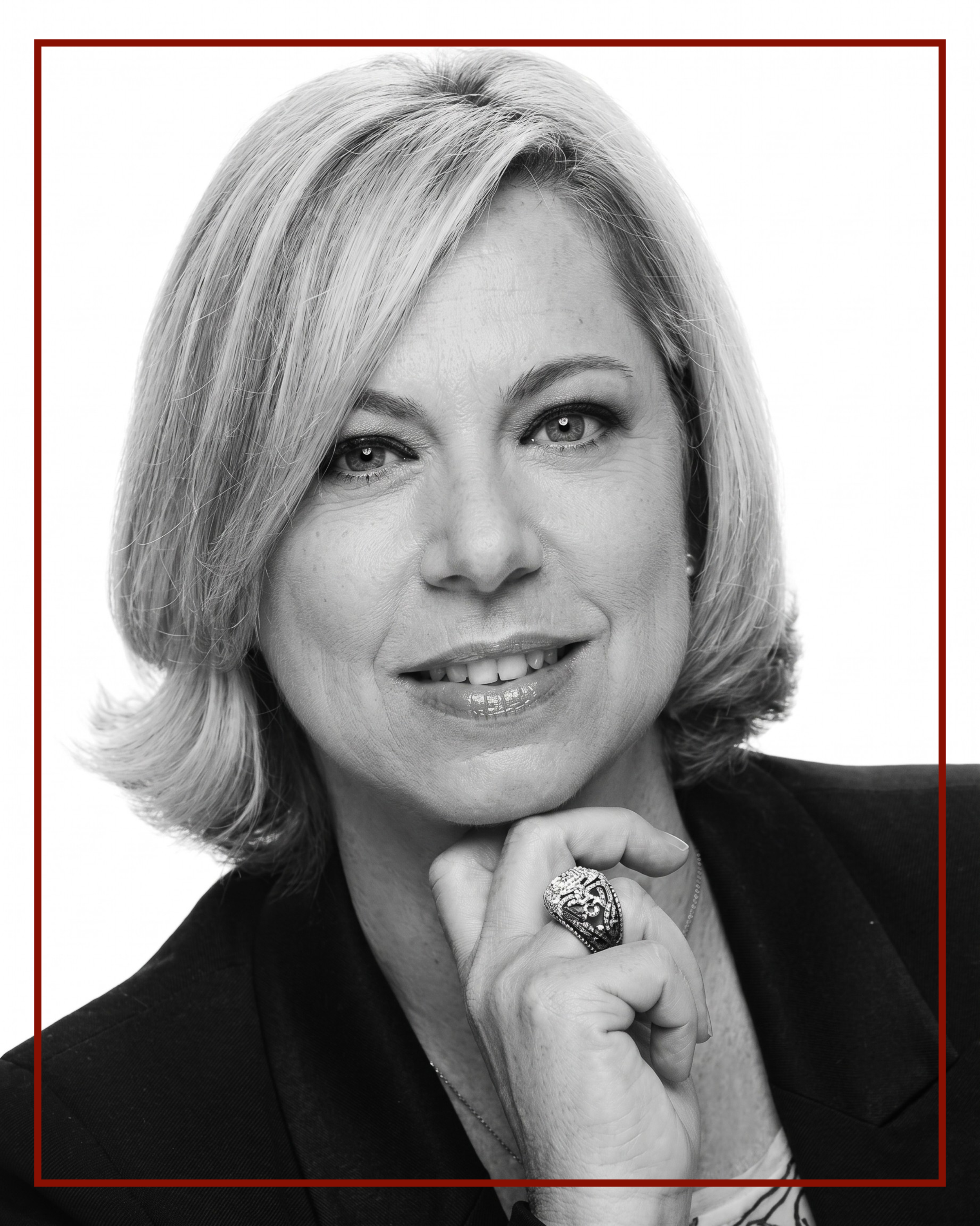 Black and white portrait of a middle-aged woman with blonde hair, smiling slightly, wearing a dark blazer, with a ring and bracelet on her right hand, and looking directly at the camera.