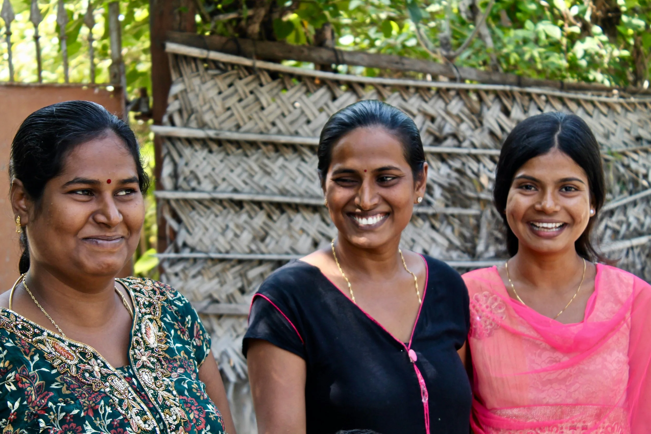 Three smiling Indian women dressed in traditional attire standing outdoors near a woven bamboo fence with green foliage in the background.
