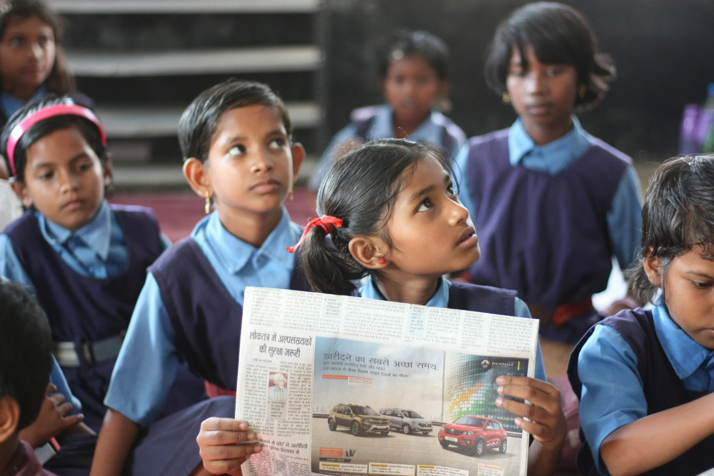 A group of young school children sitting together, with one girl reading a newspaper, in a classroom setting.