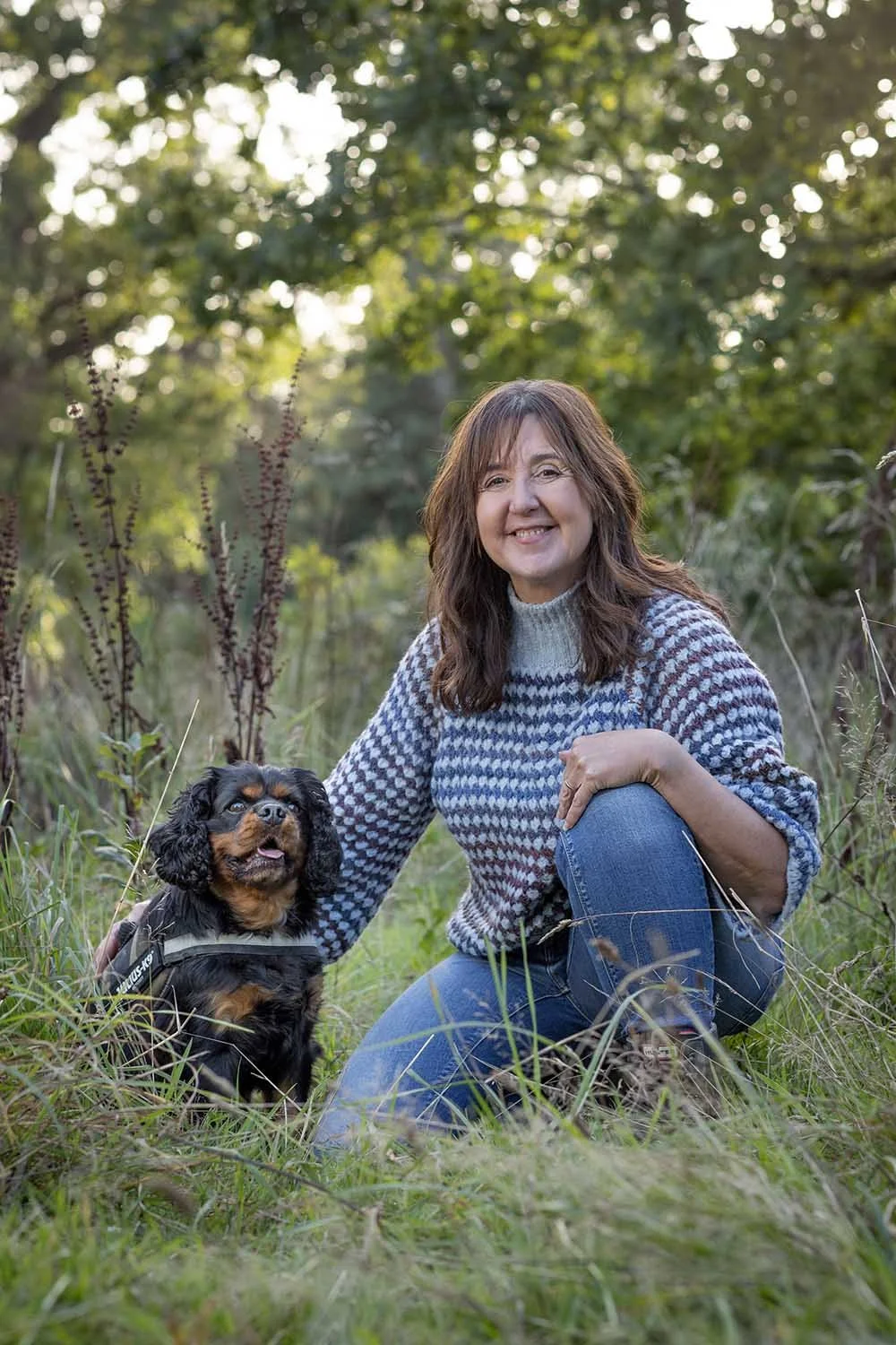 A woman kneels on grass in a forested area, smiling, with her arm around a small black and brown Cocker Spaniel dog that is sitting next to her.
