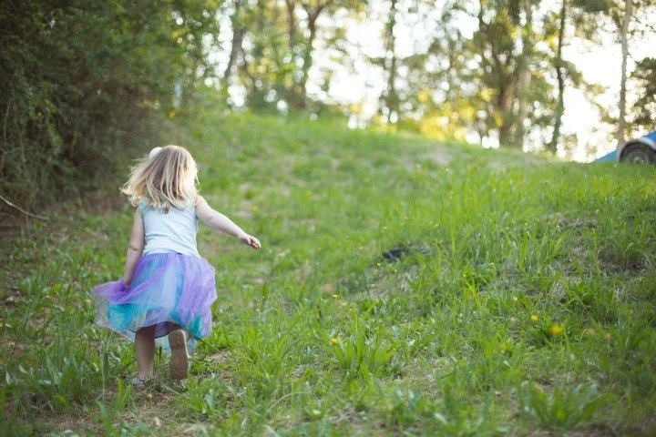 A young girl with blonde hair in a colorful tutu dress walking up a grassy hill in a wooded area.