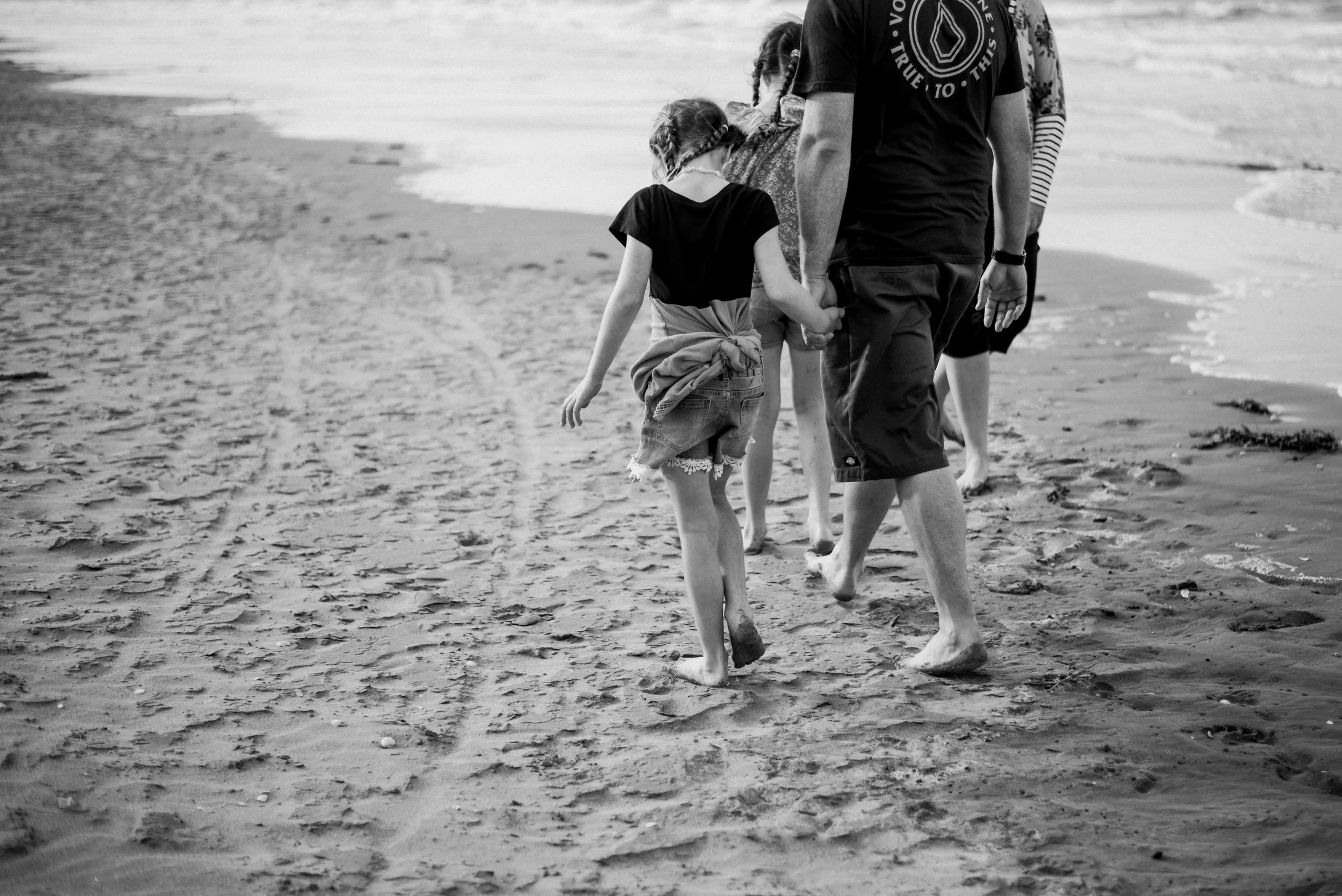 A group of people, including children and adults, holding hands while walking barefoot on a sandy beach near the water's edge.