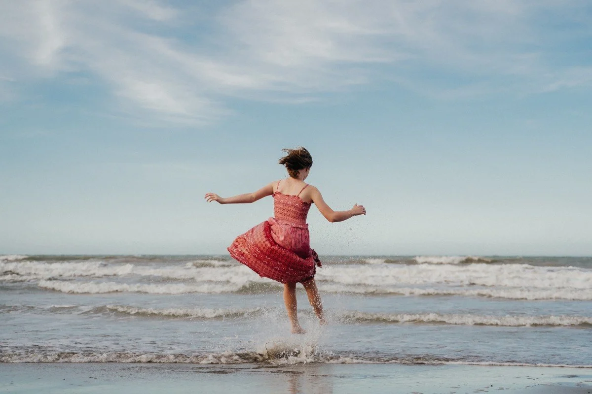 A girl in a red dress jumping and playing in shallow water at the beach with waves and a partly cloudy sky in the background.