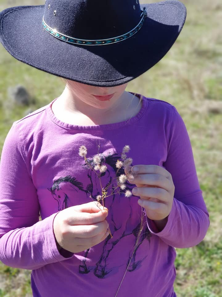 A young girl wearing a large black cowboy hat with a bejeweled band, a purple long-sleeve shirt with a horse print, is holding a small bunch of dried flowers with fluffy seed heads outdoors on a sunny day.