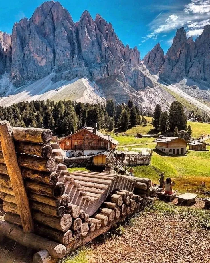 Paesaggio montano con rocce alpine e un villaggio dotato di case in legno, panchine e bambini seduti all'aperto, con una vasta area verde e alberi, sotto un cielo azzurro con nuvole.