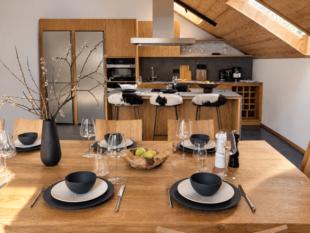 Una tavola apparecchiata con piatti, bicchieri e posate in una sala da pranzo moderna, con un ambiente cucina sullo sfondo e un grande vaso con rami decorativi al centro.
