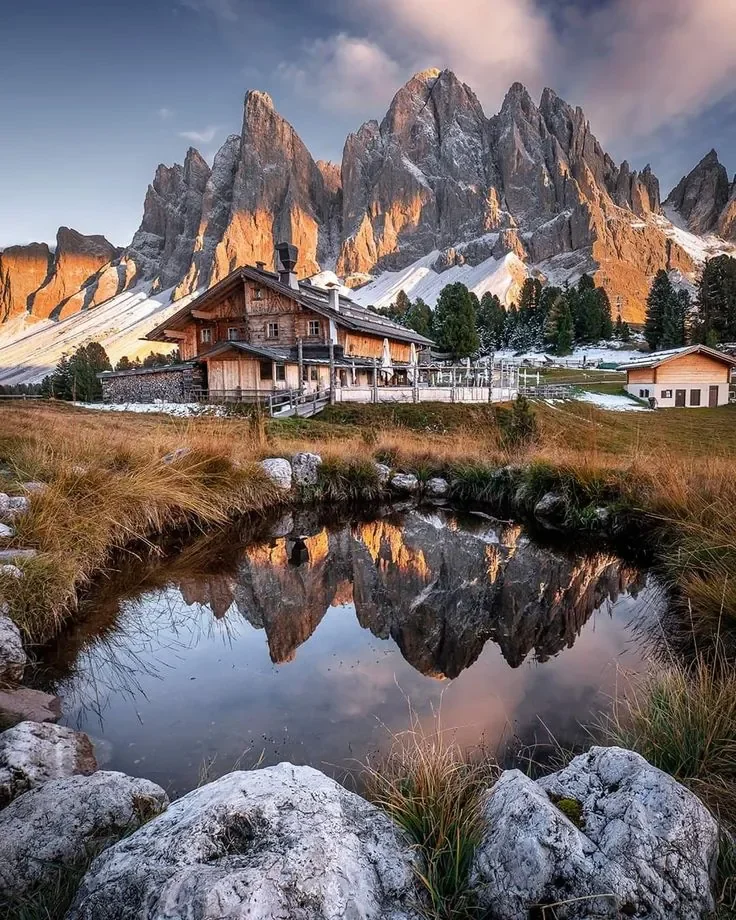 Casale di legno in montagna circondato da alberi e montagne rocciose, con un piccolo stagno riflettente il paesaggio, al tramonto.