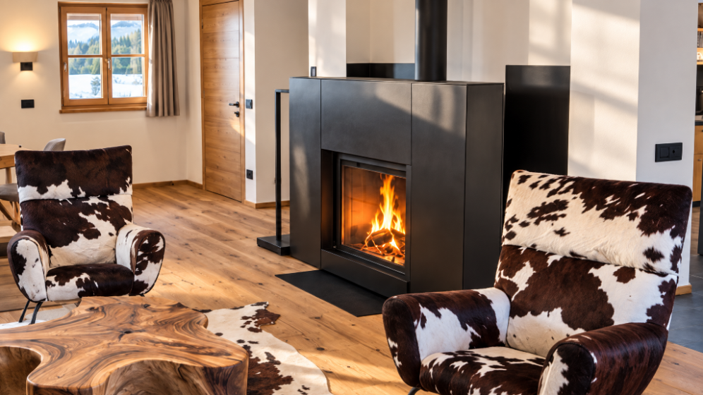 Soggiorno con camino acceso, sedie con rivestimento in pelle a motivo cowhide, tavolo in legno naturale, finestra con vista sulla natura, pareti di tonalità neutra e ambiente caldo ed accogliente.