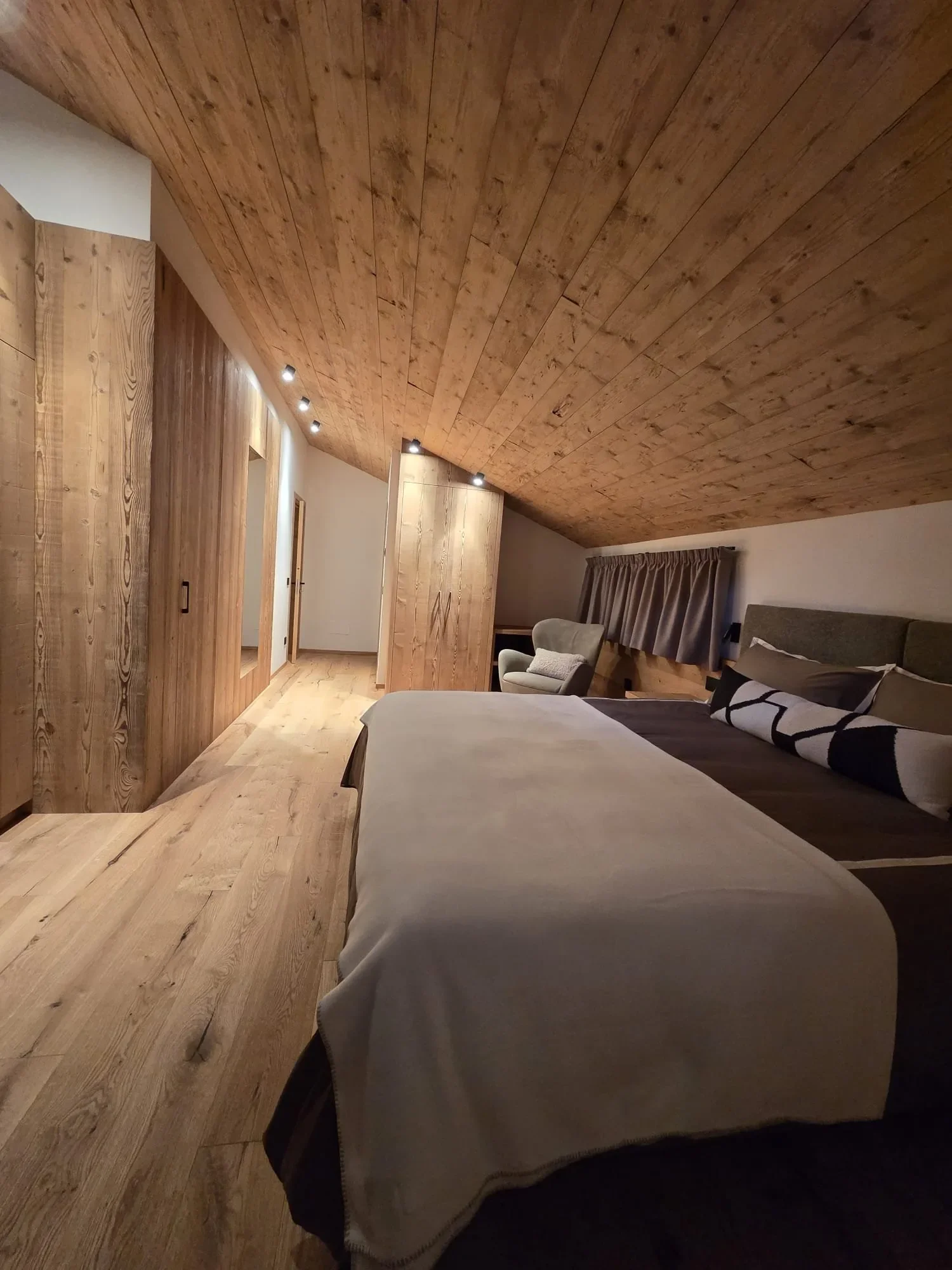 Camera da letto con pareti e soffitto in legno, letto con biancheria chiara, divano con cuscini e grande finestra con tende, soffitto con luci direzionali.