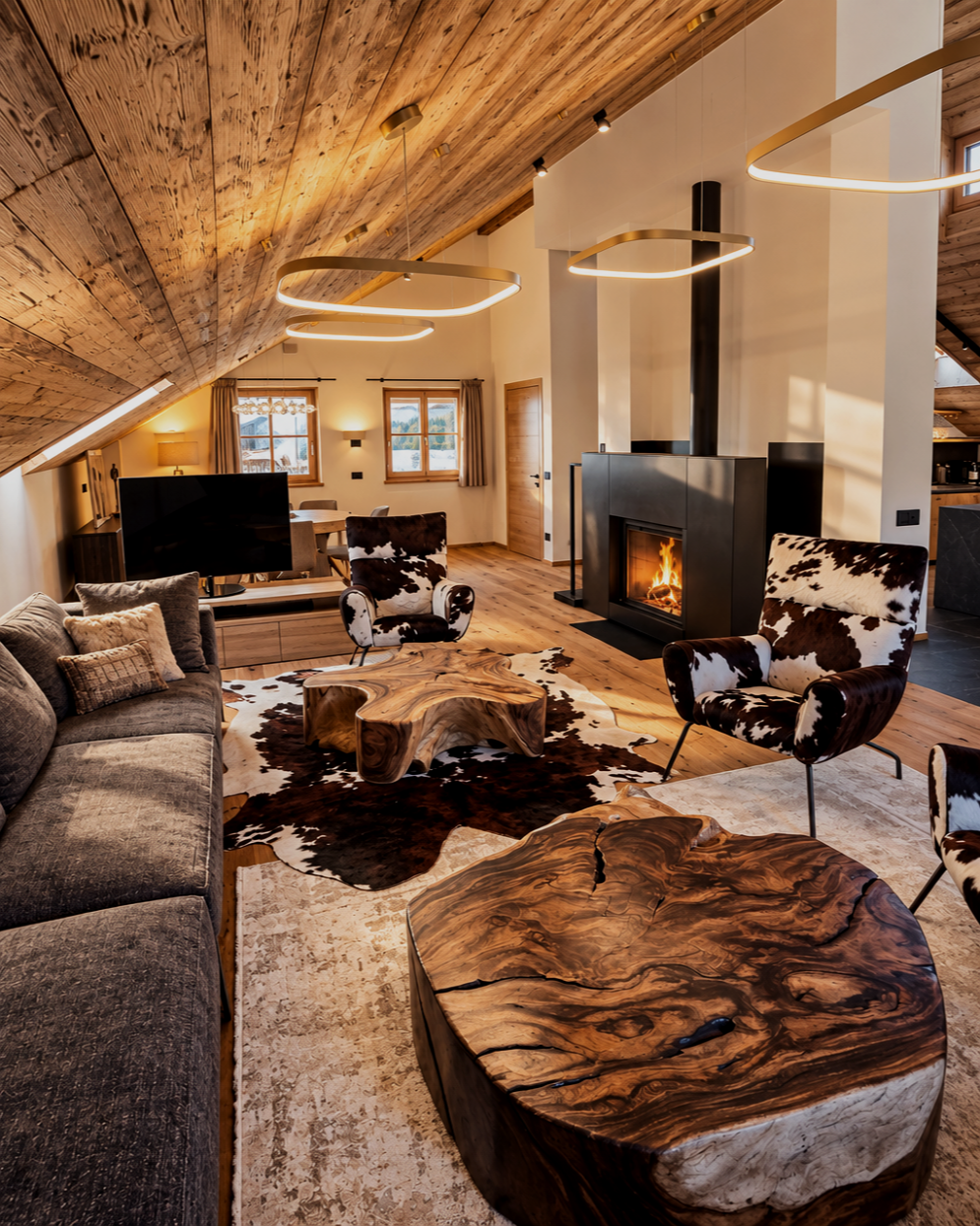Soggiorno del Chalet Gufo con soffitto in legno inclinato, camino acceso, poltrone con fantasia cowhide, tavolini in legno naturale, divano grigio e decorazioni in stile rustico.
