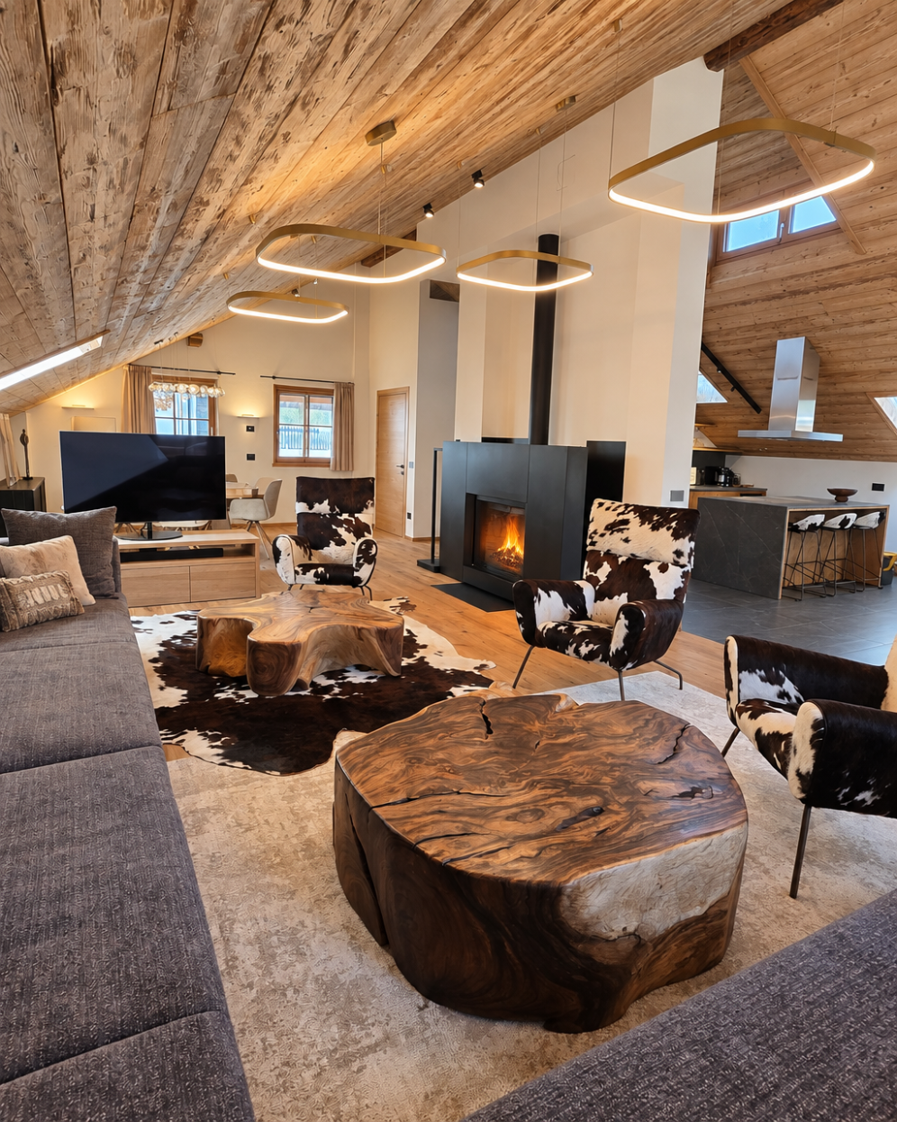 Soggiorno con divano, sedie con rivestimento in pelo di mucca, tavoli in legno massiccio, camino acceso, soffitto in legno con luci moderne, tutto al Chalet Gufo