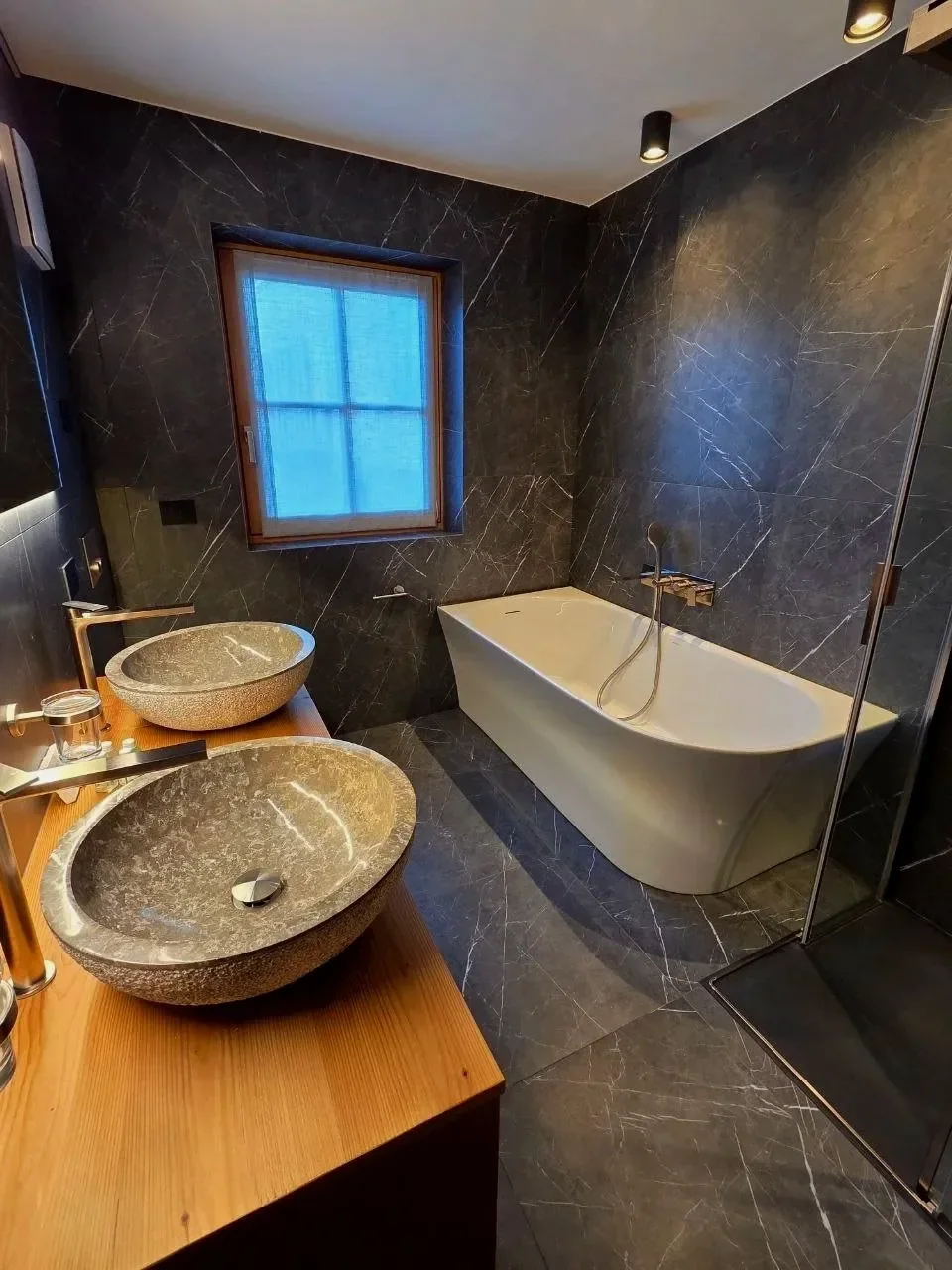 Bagno moderno con due lavabi tondi in pietra su mobile in legno, una vasca da bagno bianca, tessuti scuri in marmo sulle pareti e sul pavimento, finestra con tende blu, con luci soffuse.