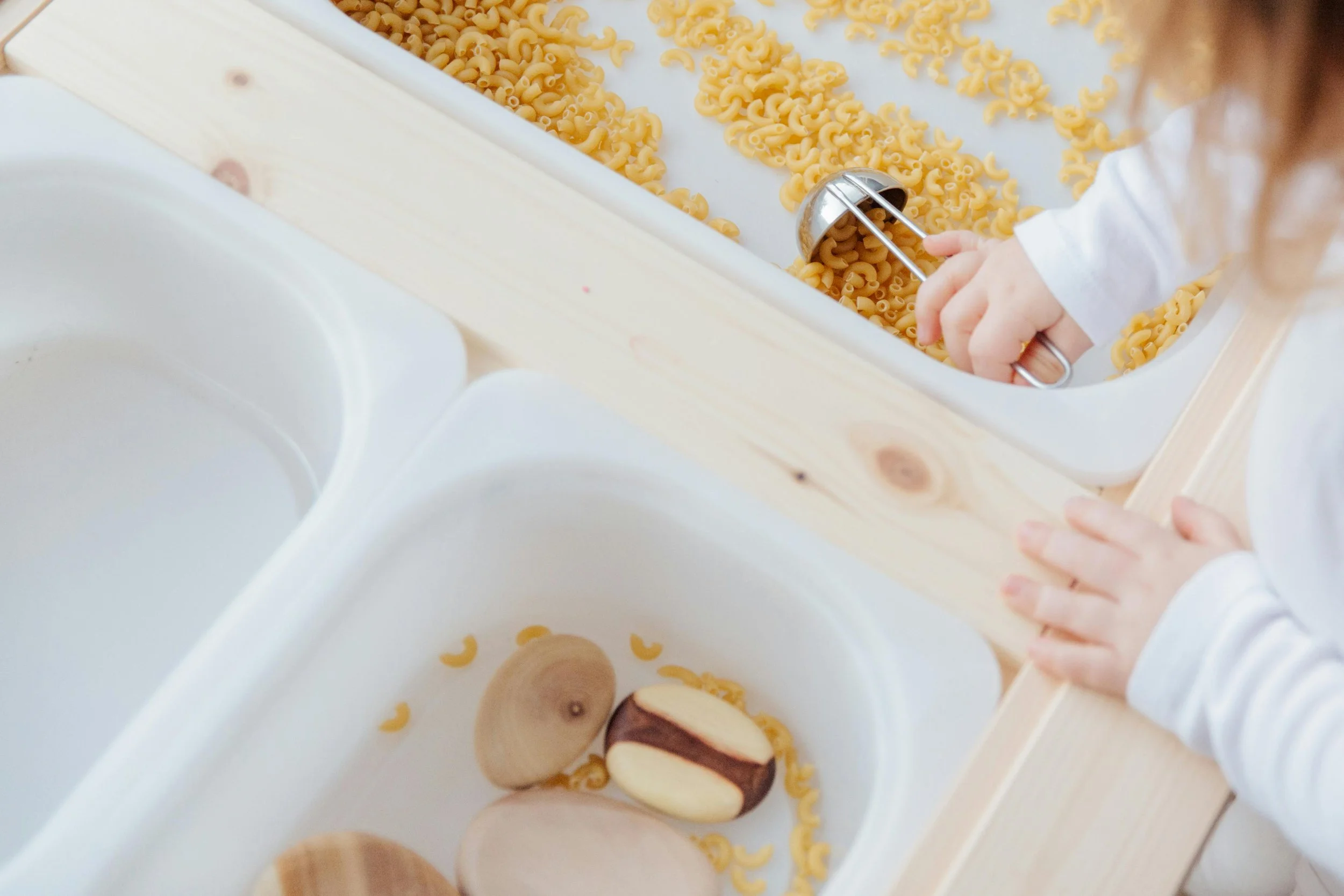 Child using a small metal scoop to transfer cooked elbow macaroni pasta onto a tray with some vanilla and chocolate sandwich cookies.